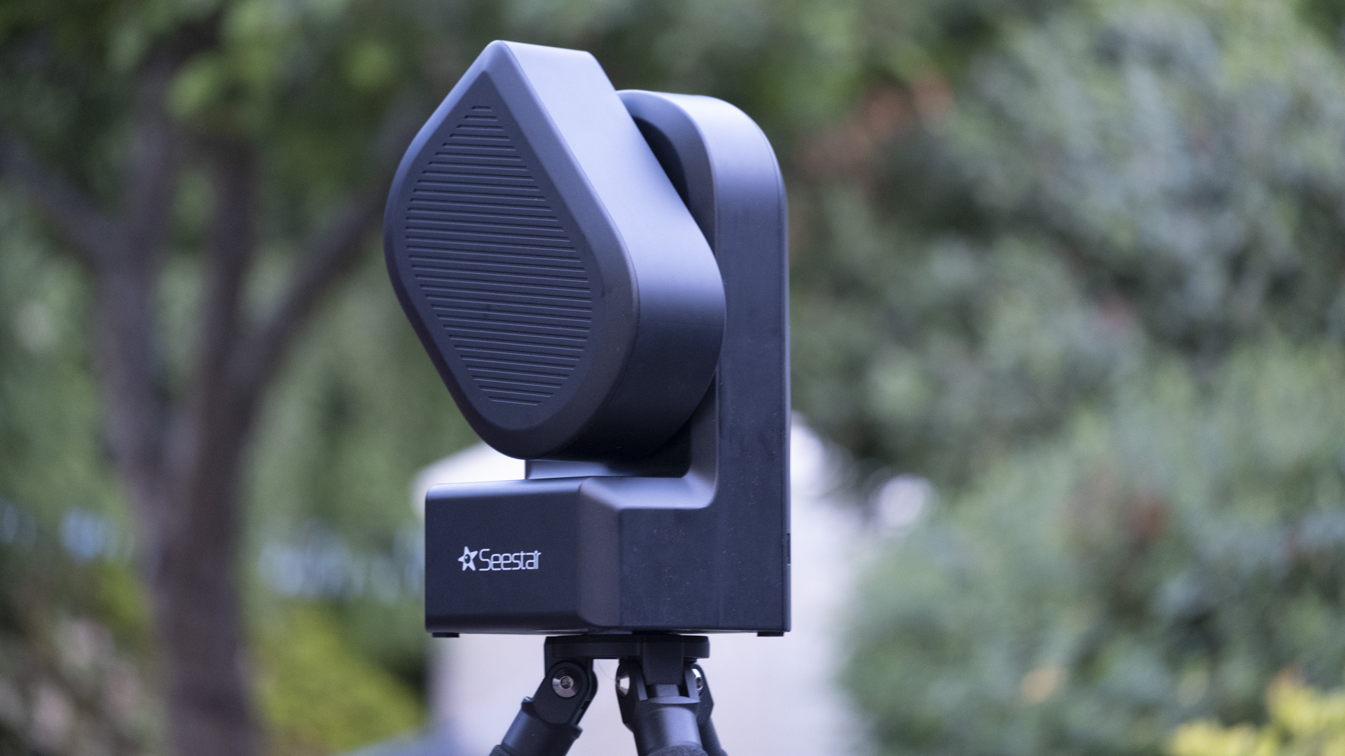 The ZWO Seestar S50 smart telescope in a garden in the daylight. In the background are trees