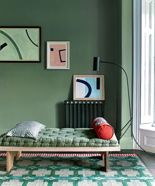Green hallway with painted green walls, daybed with wooden frame and green cushions, abstract artwork on walls in blue and pink, green patterned rug, black floor lamp beside daybed