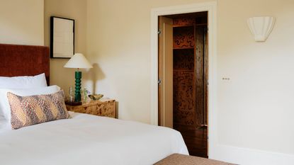 neutral bedroom with a dark red headboard, white sheets, a patchwork throw pillow, burl wood side table and a shell wall sconce