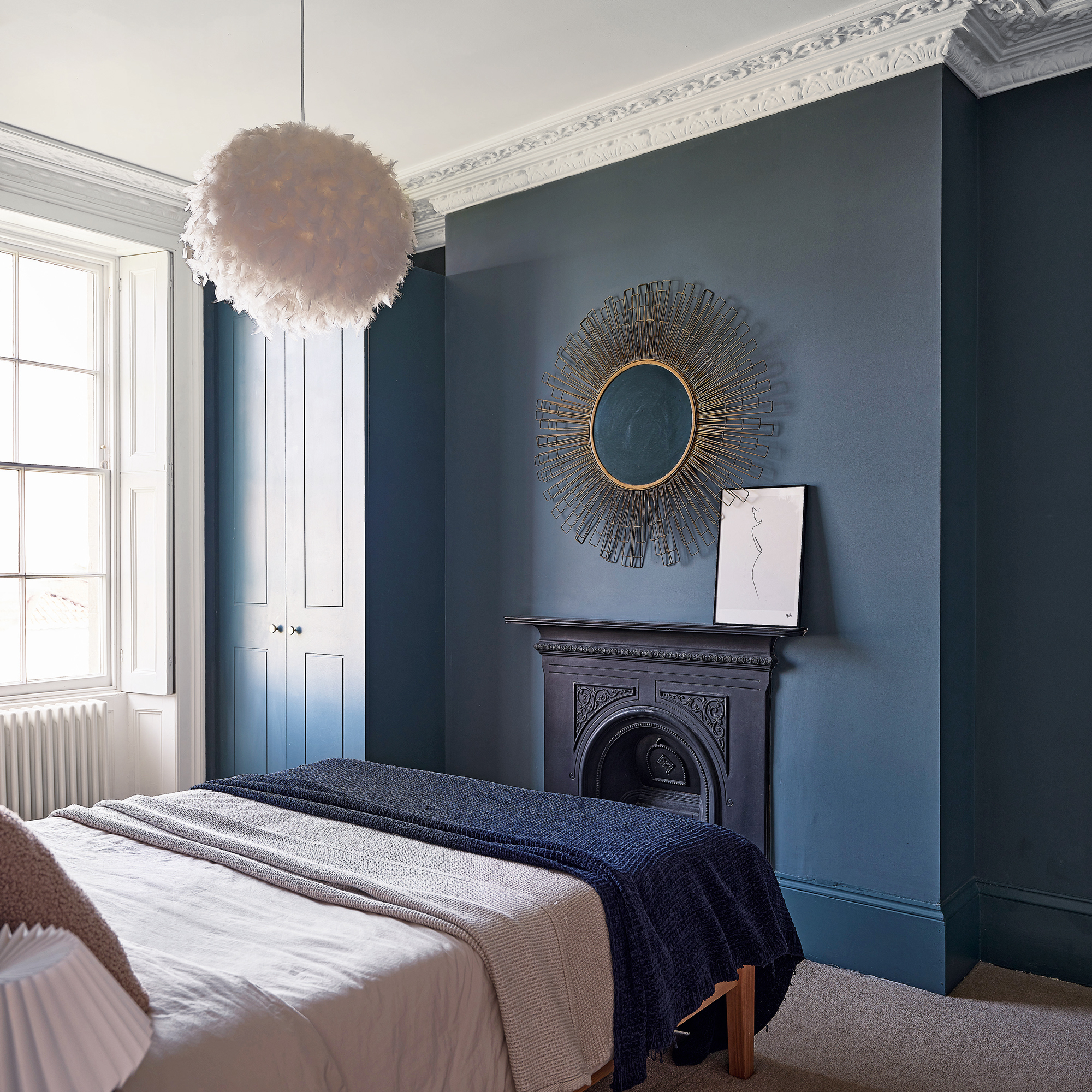a navy blue bedroom with colour-drenched fitted wardrobe, a starburst mirror, fireplace and feather pendant light
