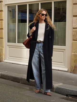 Fashion influencer Anouk Yve with long, curly hair on the street in Europe wearing belted wide-leg jeans, a cream knit sweater, a long wool coat, sunglasses, and mules and carrying a brown tote.