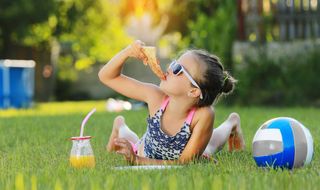 A smiling child eats a slice of pizza in the sunshine while reclining on a picnic rug on a summer's day