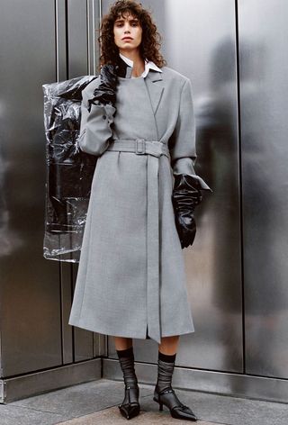 One of the best jacket coat brands, Mango, is shown in an image of a model wearing a gray coat with leather gloves, black sheer socks, and black kitten heels