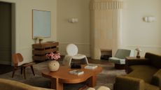 A beautiful living room with a tiled fireplace, a white pendant lamp, curved wall sconces, a wood sideboard, blue art, a wooden chair, a white sofa chair, an armless blue chair, a deep green sofa, a beige sofa, and a wooden coffee table with a vase of flowers and books