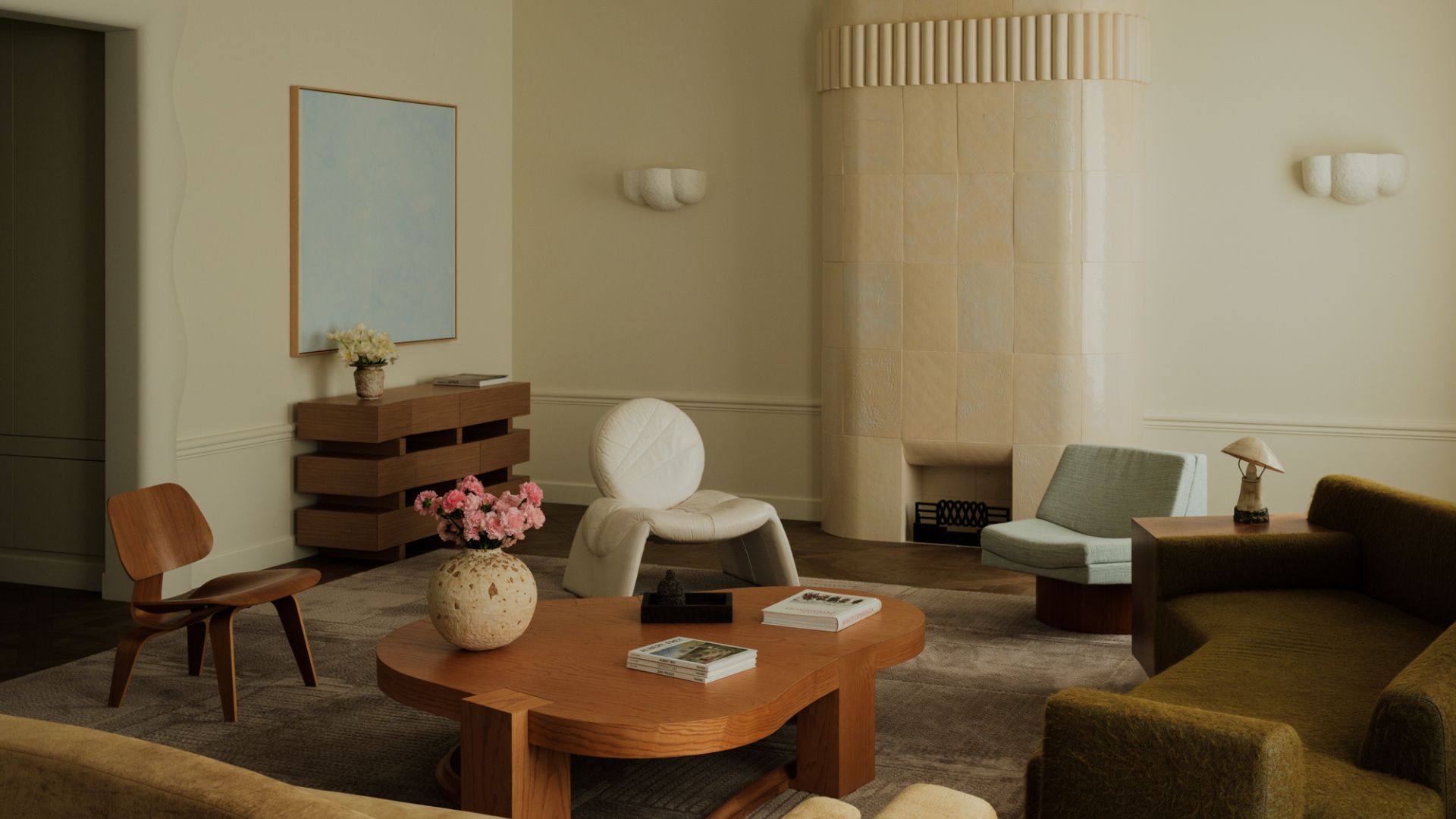 A beautiful living room with a tiled fireplace, a white pendant lamp, curved wall sconces, a wood sideboard, blue art, a wooden chair, a white sofa chair, an armless blue chair, a deep green sofa, a beige sofa, and a wooden coffee table with a vase of flowers and books