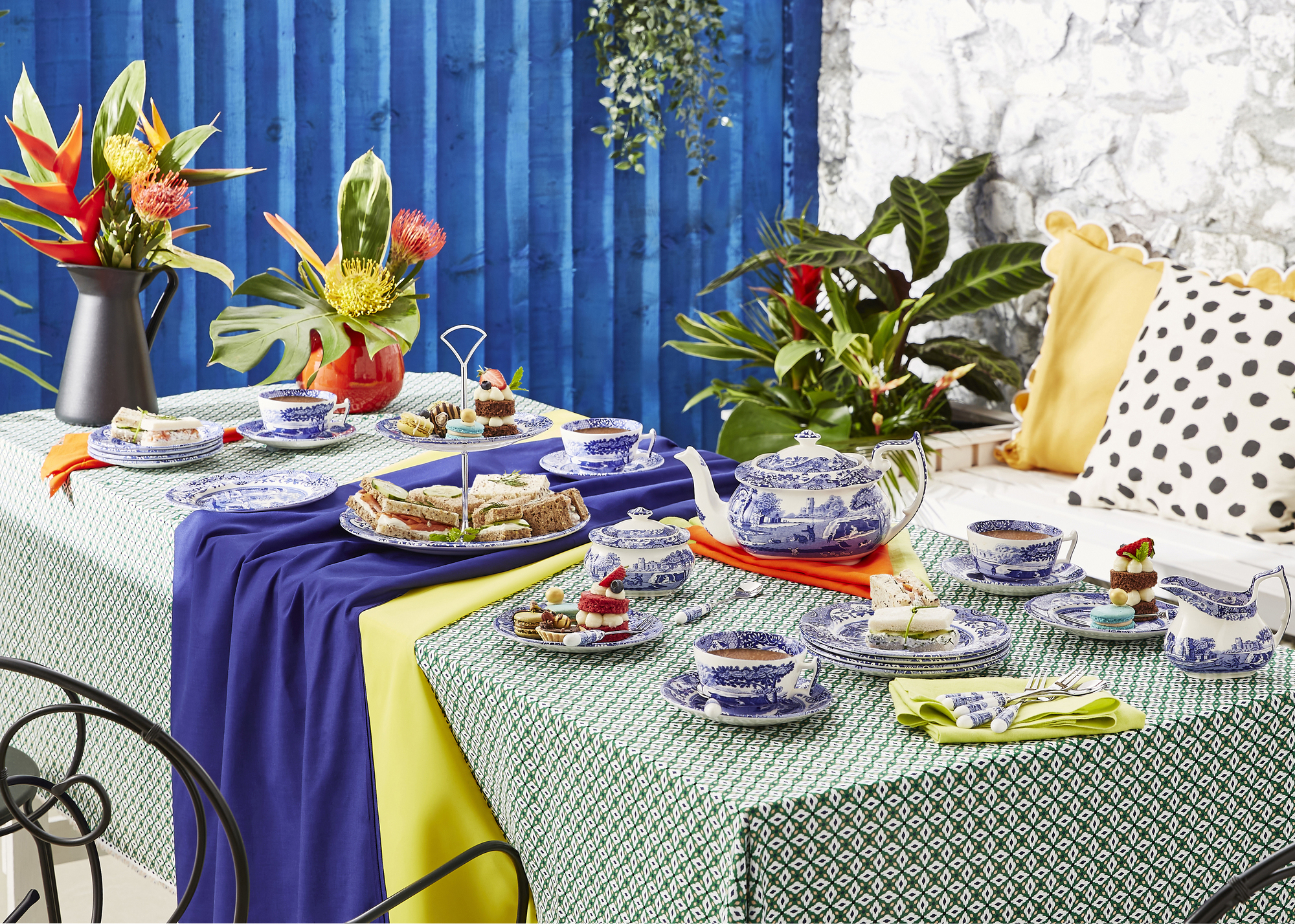 An outdoor table set for afternoon tea with a green patterned tablecloth, blue china tableware, and colorful vases of flowers