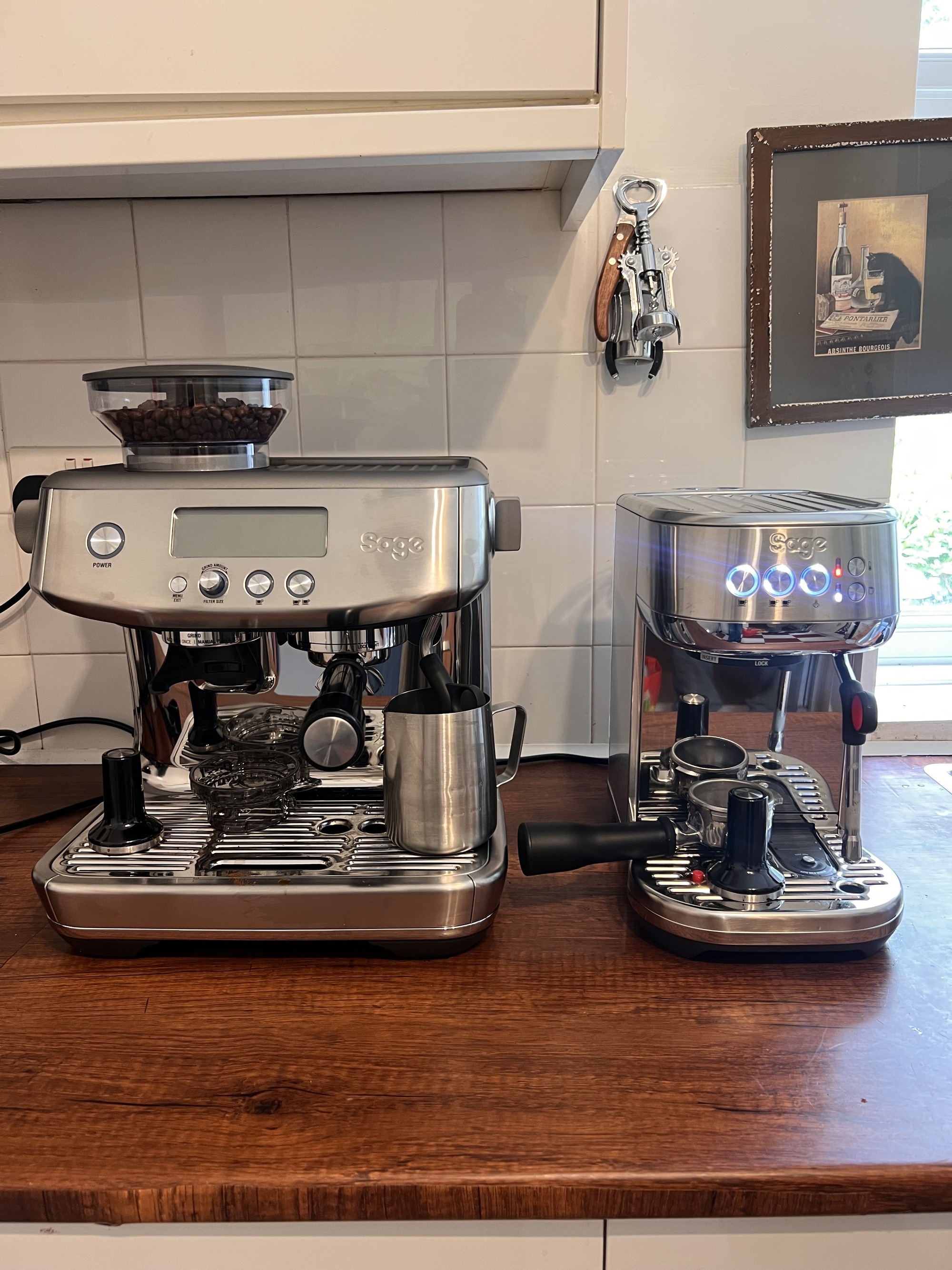 Image of a silver Sage Bambino Plus next to a silver Sage Barista Plus  on a wooden counter in a white kitchen.