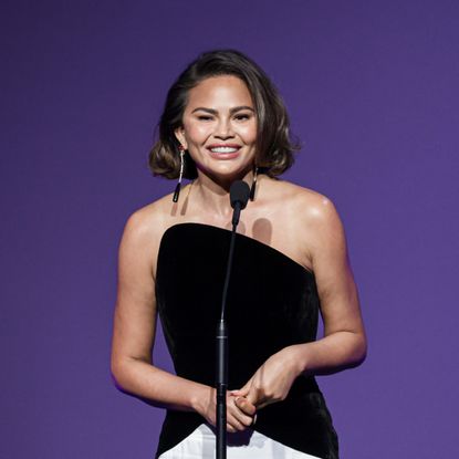 chrissy teigen speaks on stage wearing a black and white gown