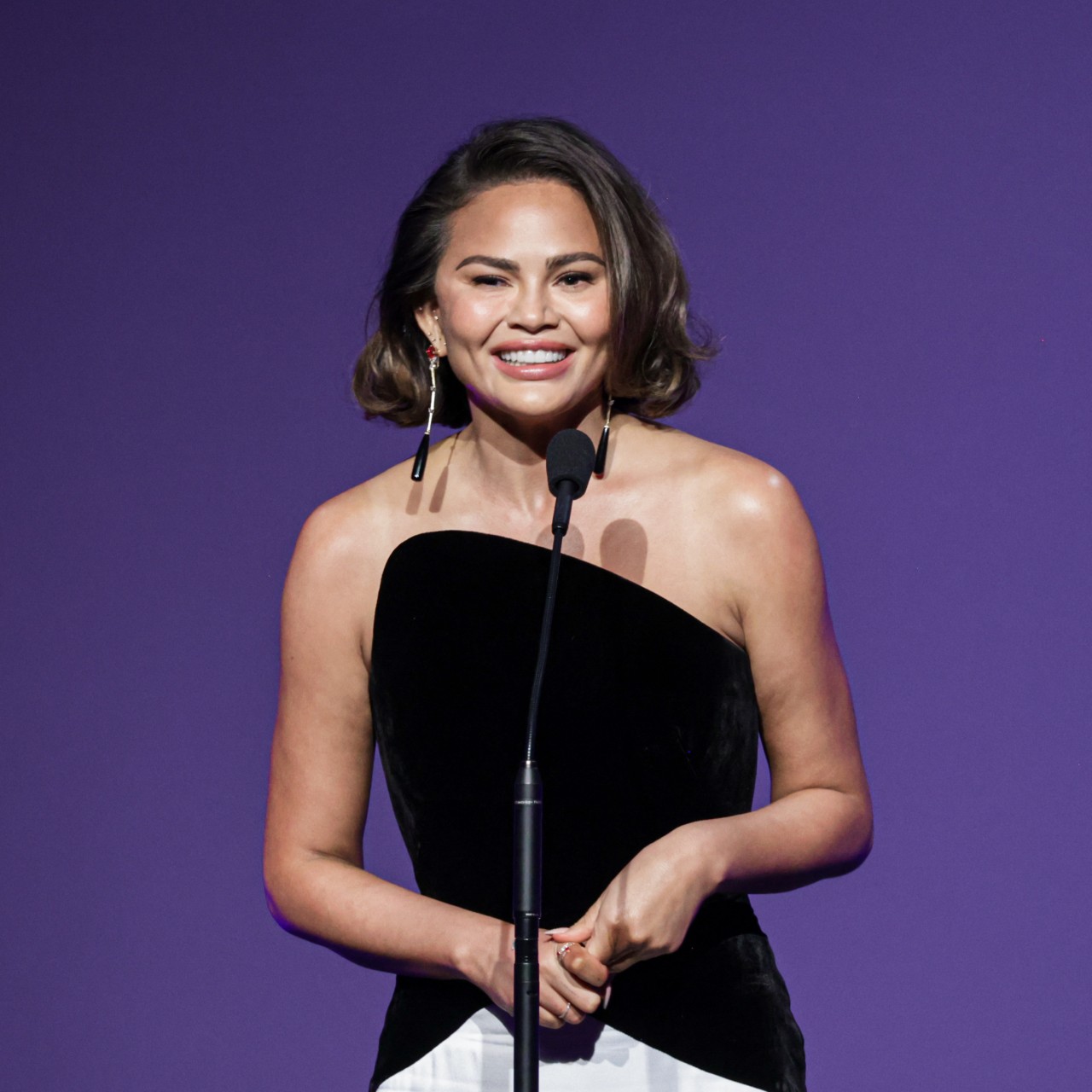 chrissy teigen speaks on stage wearing a black and white gown