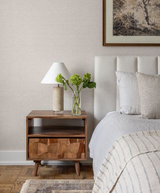 Bedroom with wood nightstand and part of bed dressed with neutral linens in front of neutral wall with neutral patterned area rug beneath