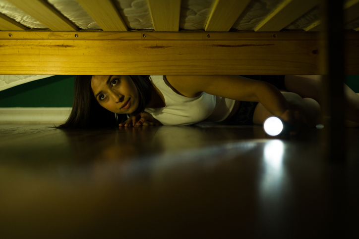 A woman looking under the bed with a flashlight.