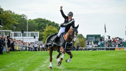 Gemma Stevens, a rider wearing a black coat and a sash, raises an arm on her Burghley lap of honour with Chilli Knight, a chestnut horse, who wears the winners&rsquo; rug