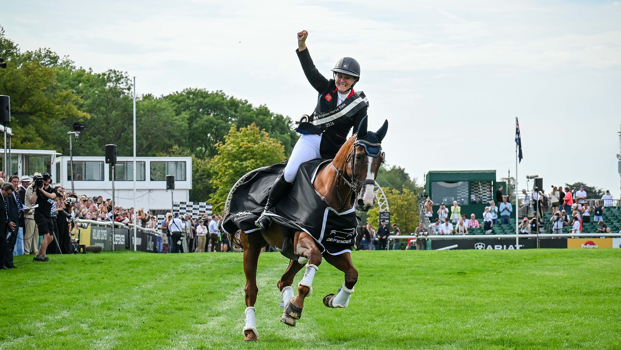 Gemma Stevens, a rider wearing a black coat and a sash, raises an arm on her Burghley lap of honour with Chilli Knight, a chestnut horse, who wears the winners&rsquo; rug