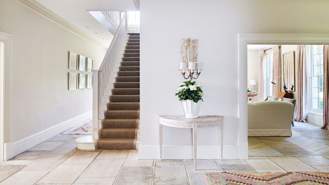 A bright white entryway with a brown stair runner, white table with flowers in a white vase, and a living room to the right with large windows