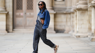 Emilie Joseph @in_fashionwetrust wears black large butterfly sunglasses, a navy blue denim shoulder-pads / cropped denim jacket, a black faded denim baggy overall / dungarees/ jumpsuit by John Galliano, brown metallic leather with white details sneakers from Valentino, during a street style fashion photo session, on October 16, 2022 in Paris, France. (