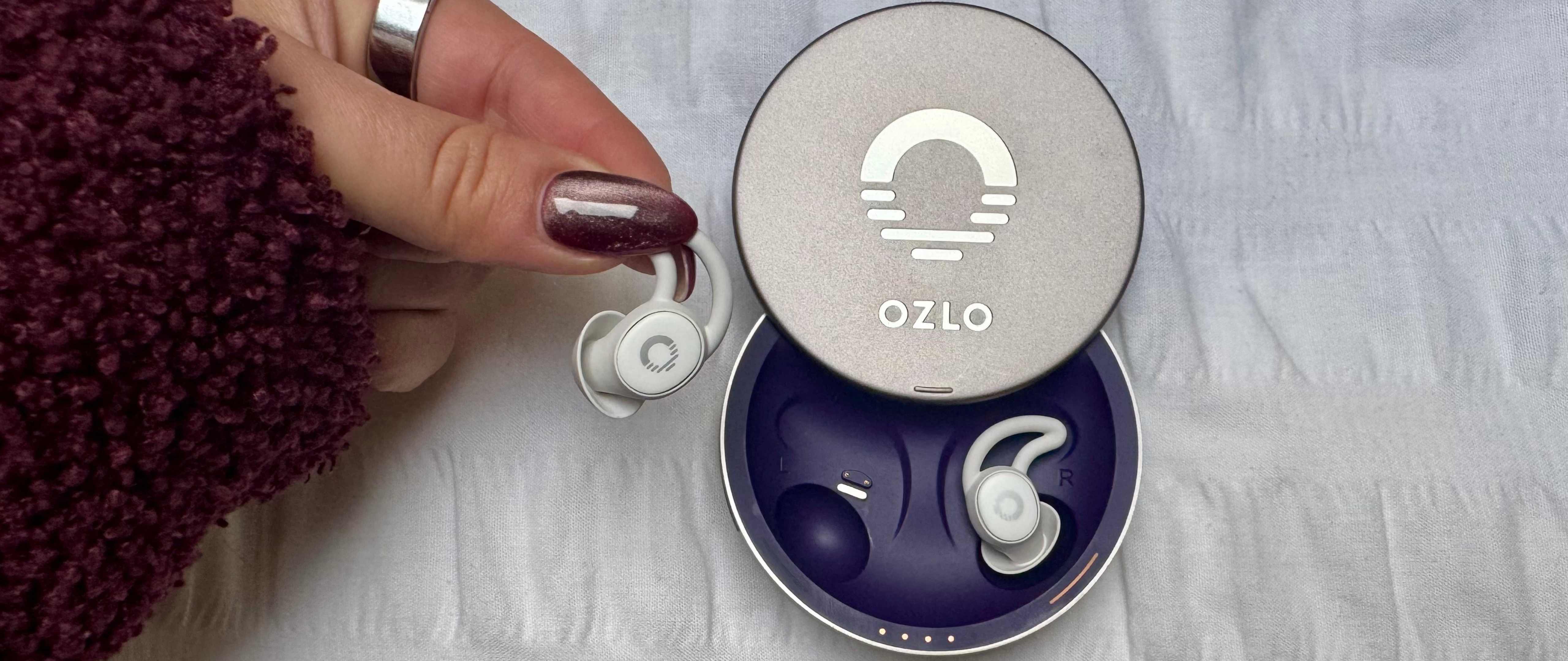 A close-up shot shows a hand holding a white Ozlo Sleepbud next to its open, circular, silver-grey charging case. The case lid features the Ozlo logo and the word &quot;OZLO.&quot; The Sleepbuds have a small, earhook-like design. The background is a white, slightly textured fabric.