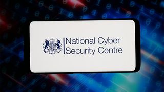 Insignia of the UK's National Cyber Security Centre (NCSC) pictured on a smartphone screen with blurred blue, green, and orange coloring in background.