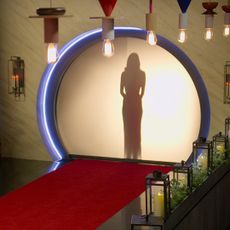 A woman stands behind a backlit screen, seen from a room decorated with a red carpet, handing Edison bulbs, and candle displays, in 'Love is Blind' season 7.