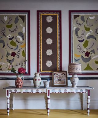 Colorful hallway with painted, patterned console table, three large artwork designs on walls above, console decorated with table lamp, ornaments