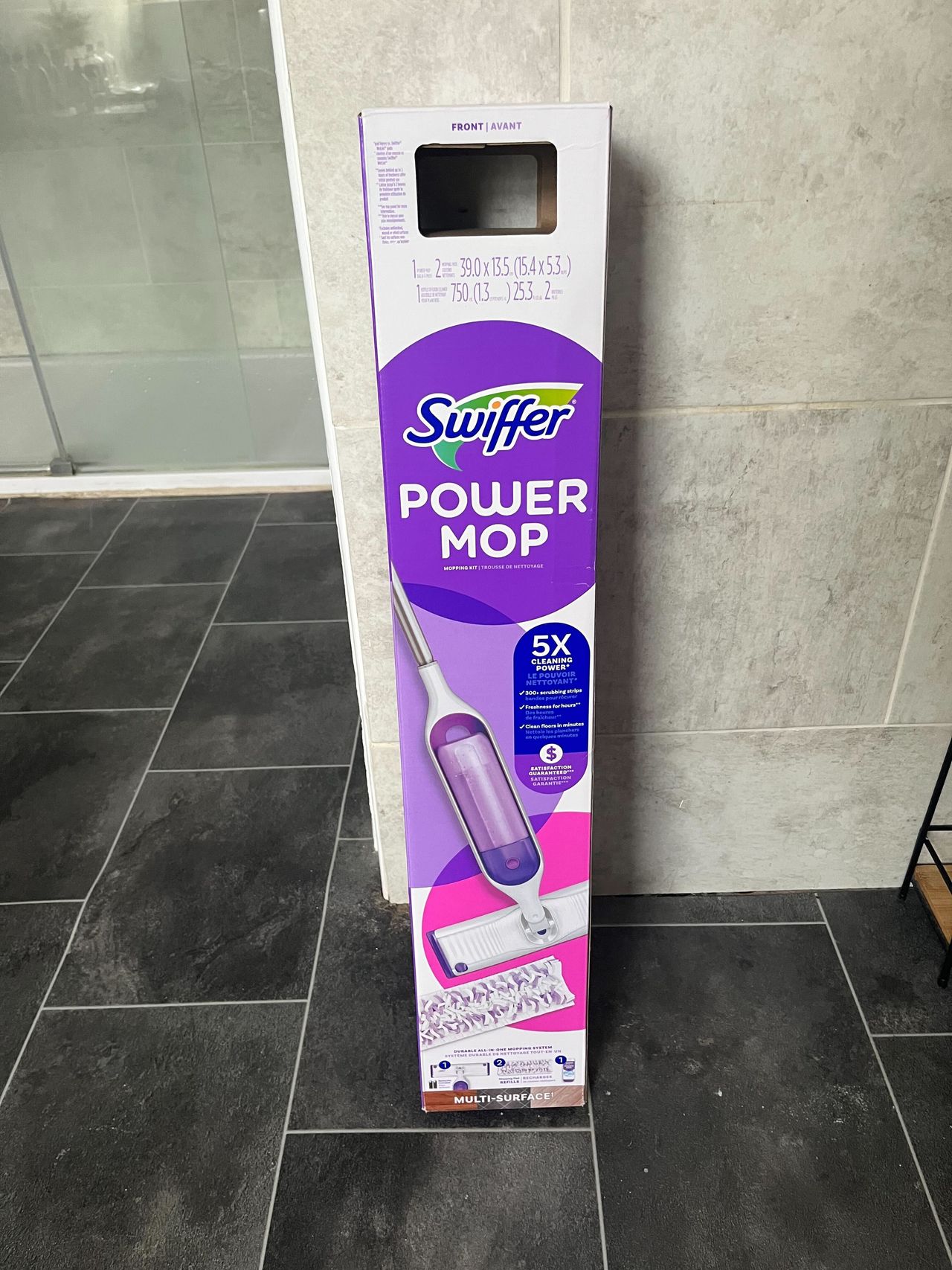 a purple and white Swiffer power mop in carboard packaging leaning against a tile wall