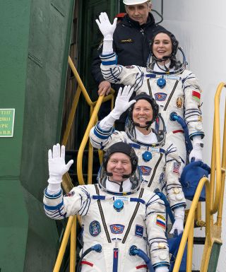 three astronauts on a staircase outside a rocket, in spacesuits, waving and smiling