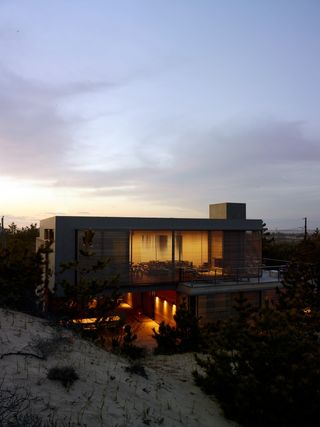 The beach house photographed at a sunset. A two-floor house, with panoramic windows, sits surrounded by sand and different plants.