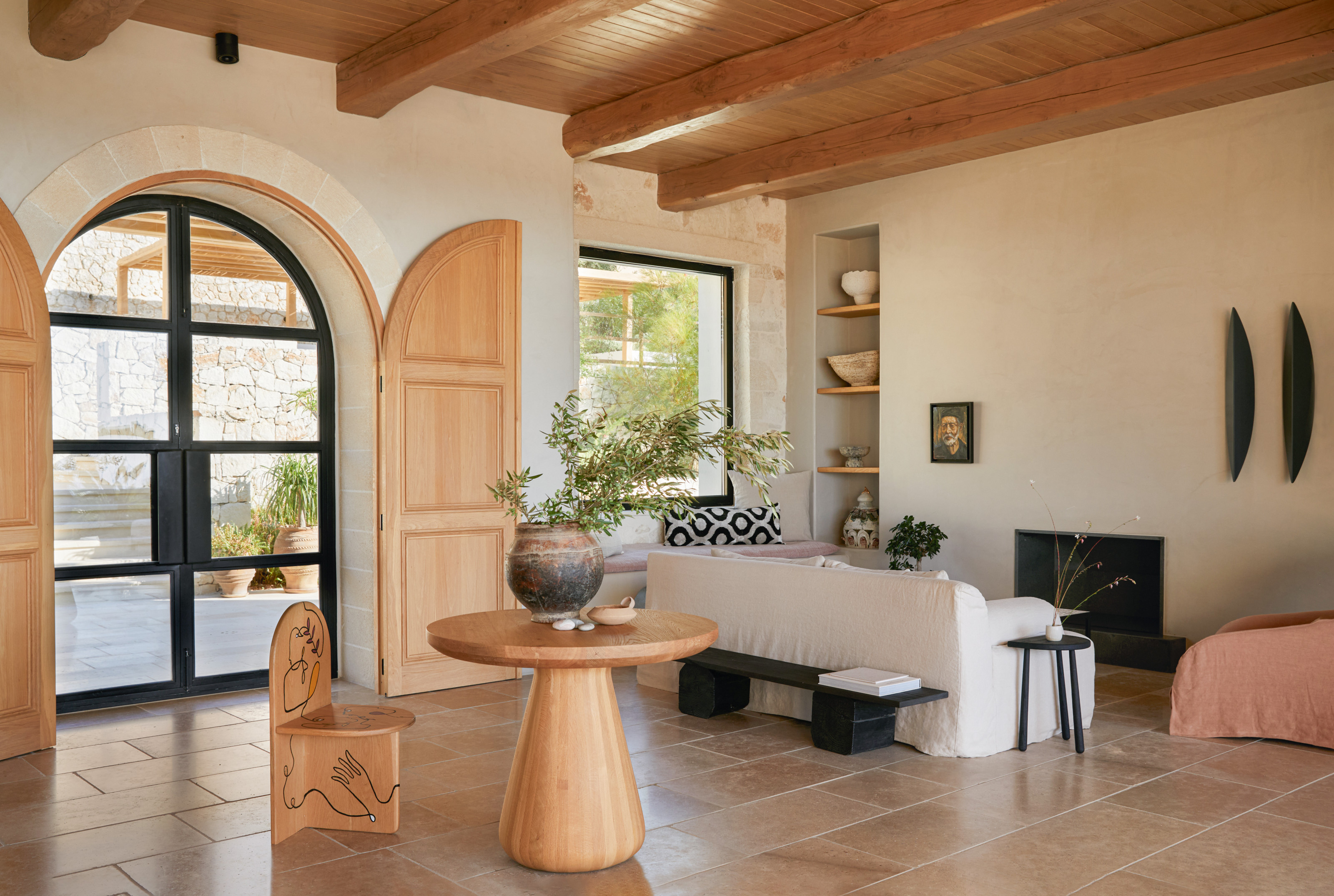 Hallway with warm off-white walls, exposed ceiling beams, stone floor, arched glazed door, round wood entrance table and sculptural wood chair
