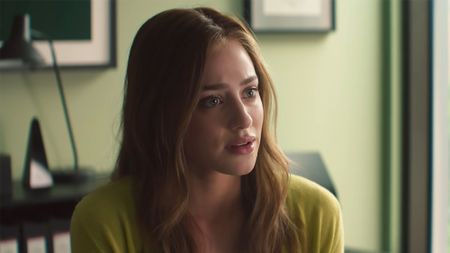 Lili Reinhart looks concerned while sitting at Carlsen's desk in The Love Hypothesis. Her hair is down and she's wearing a cardigan.