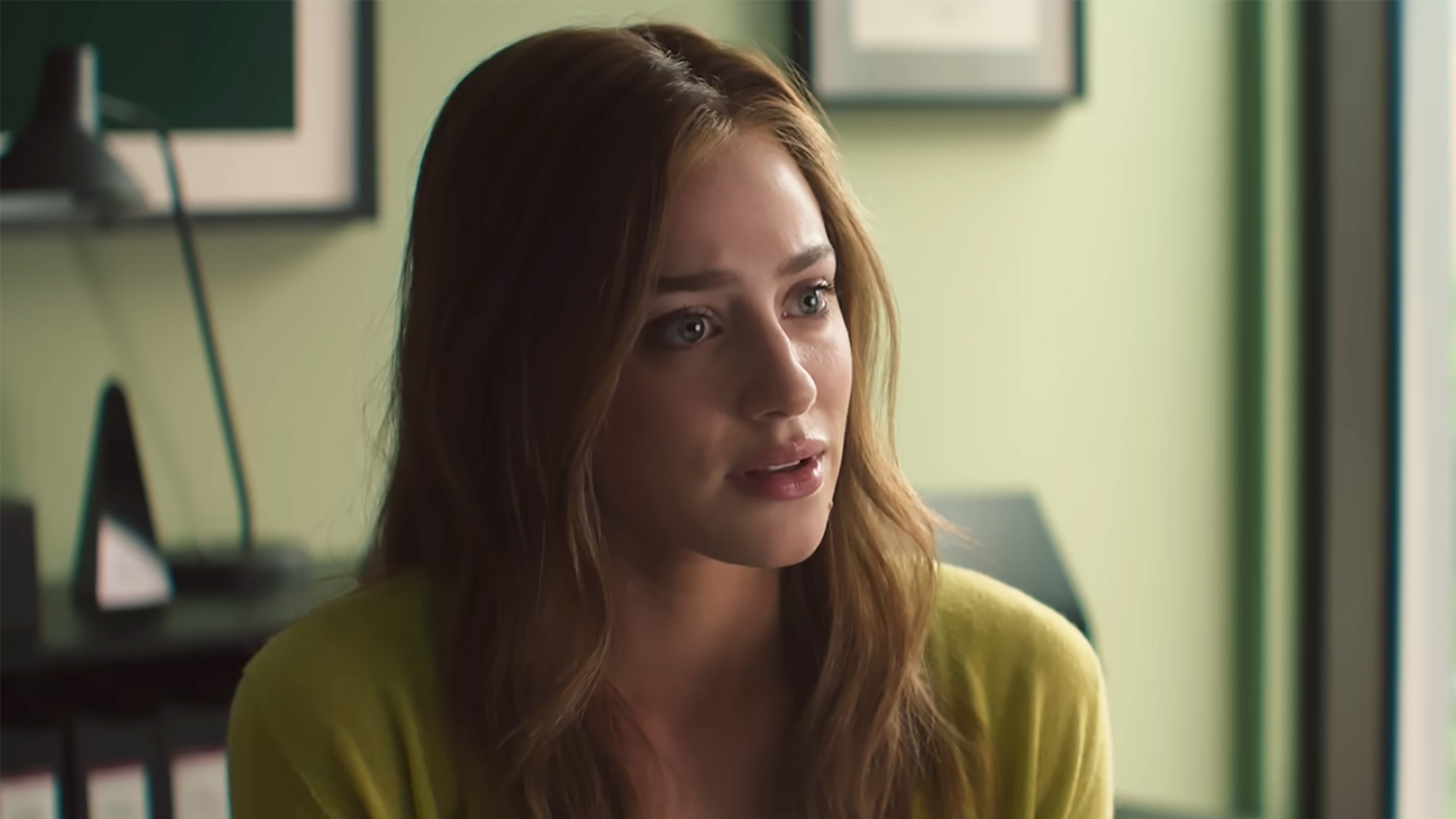 Lili Reinhart looks concerned while sitting at Carlsen's desk in The Love Hypothesis. Her hair is down and she's wearing a cardigan.