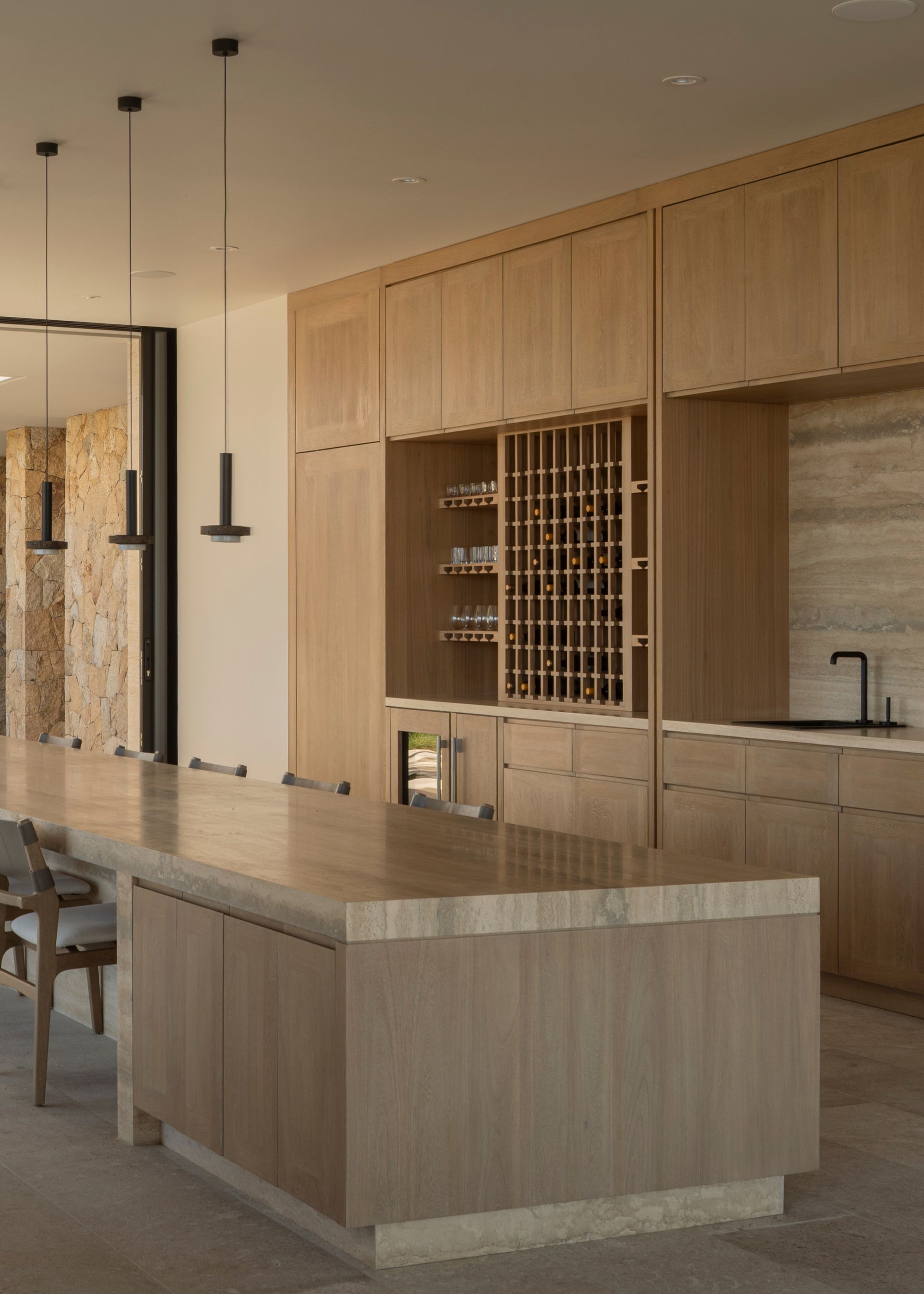 A large modern kitchen with an open wine wall, closed wood cabinets, a long kitchen island, and a row of gray chairs