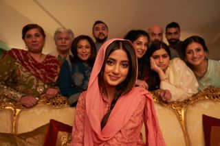 Maymouna wearing a sari and sitting on a chair as her family eagerly stand behind her with a mixture of emotions.