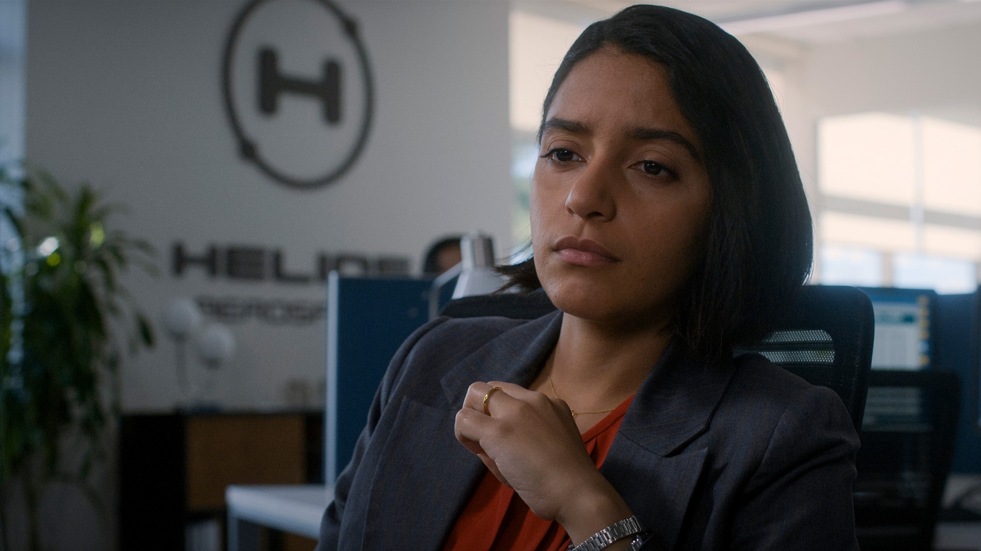 Close up of a woman sitting down. She is wearing a business suit with an orange shirt underneath. She has shoulder-length dark hair. She has a serious expression on her face as she contemplates the situation. In the office background there is a large H in a circle with the text 'Helios' underneath.