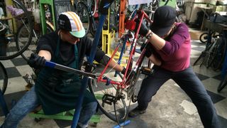 Two people wearing caps are working together to bend a steel frame back into place. Both have a wide stance, and the person on the right shoulders the front of the frame while the person on the left uses a tool to manipulate the rear of the frame