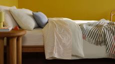 Assorted Brooklinen bedding, including striped sheets, a white duvet, and patterned throw blanket, styled on a bed in a yellow-walled bedroom with wooden furniture.