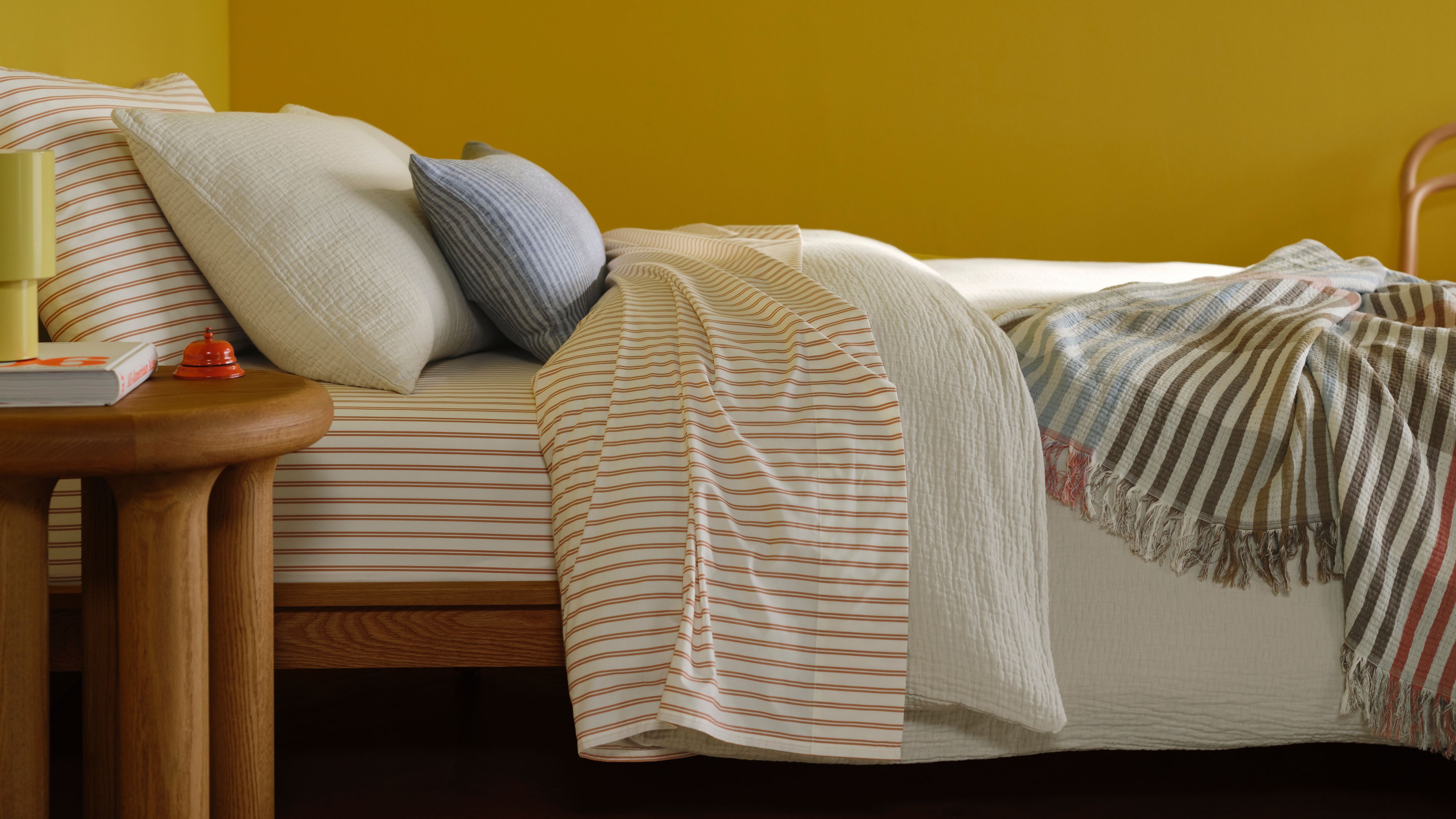 Assorted Brooklinen bedding, including striped sheets, a white duvet, and patterned throw blanket, styled on a bed in a yellow-walled bedroom with wooden furniture.