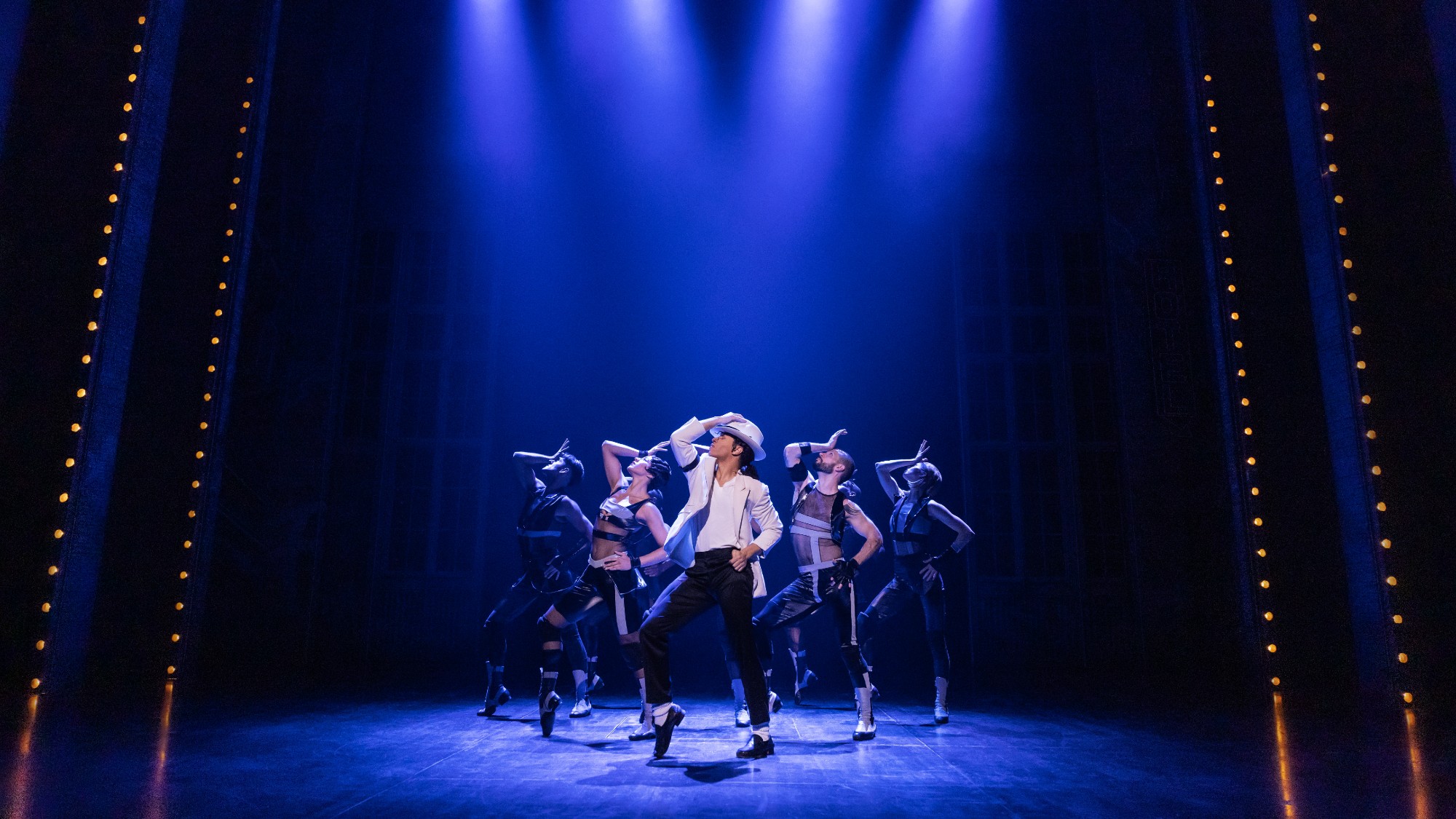 the company of the Broadway cast of MJ: The Musical striking one of Michael Jackson's signature poses. Jackson is dressed in a white hat and shirt and black pants