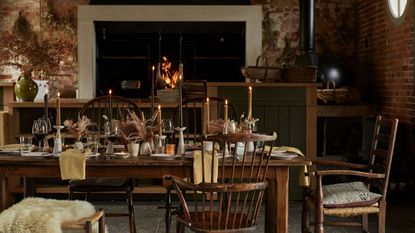 A cozy kitchen dining area with a large open fire in the background, set into an exposed brick wall. In the foreground is a large wooden dining table with wooden chairs and fur throws, set with taper candles, flowers, wine glasses, and napkins.