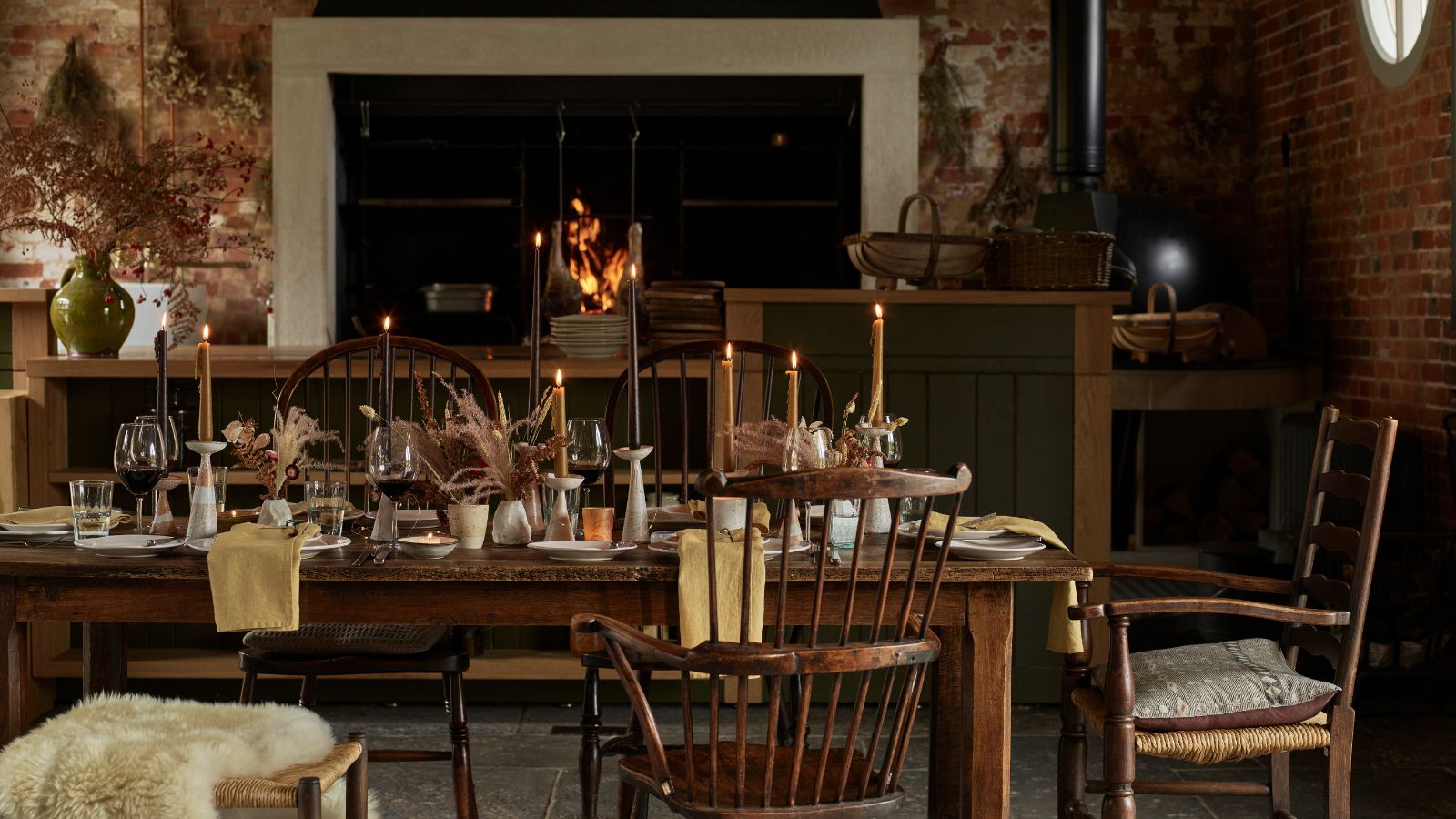 A cozy kitchen dining area with a large open fire in the background, set into an exposed brick wall. In the foreground is a large wooden dining table with wooden chairs and fur throws, set with taper candles, flowers, wine glasses, and napkins.