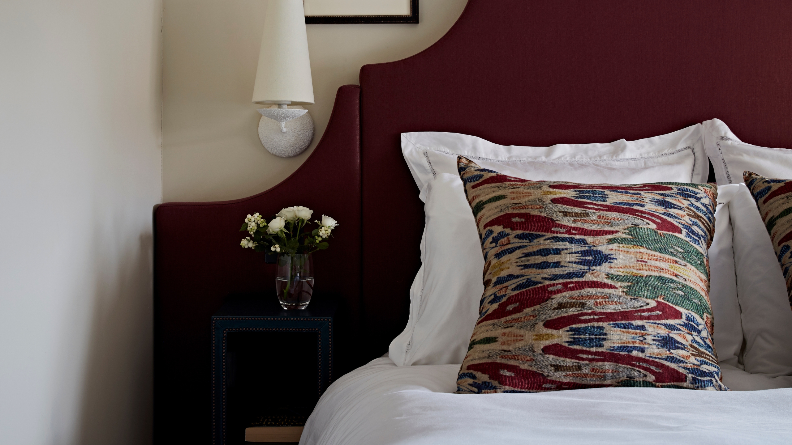 Burgundy bed with white bedding and patterned cushion, with a dark wood bedside table with small glass vase filled with white roses, and a white sconce lamp above