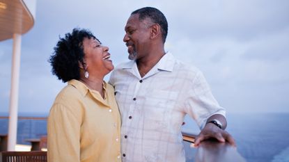 A Baby Boomer couple laugh and smile at each other while standing on the deck of a boat.