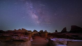 Shooting stars are pictured as lines streaking across the glowing band of the Milky Way, which is visible tumbling vertically towards the horizon. Stone ruins are visible below and a soft glow is detectable near the horizon.