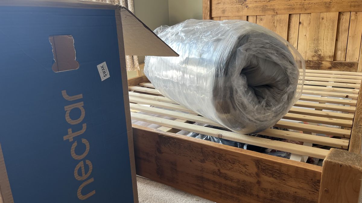 nectar classic hybrid mattress tightly rolled up in plastic on wooden bed frame next to cardboard delivery box