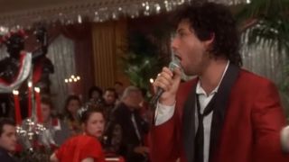 Adam Sandler singing into a microphone with a red jacket on in The Wedding Singer