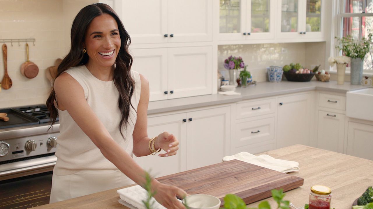 Meghan Markle wearing a white sleeveless tank and pants cooking in a kitchen with white cabinets and laughing 