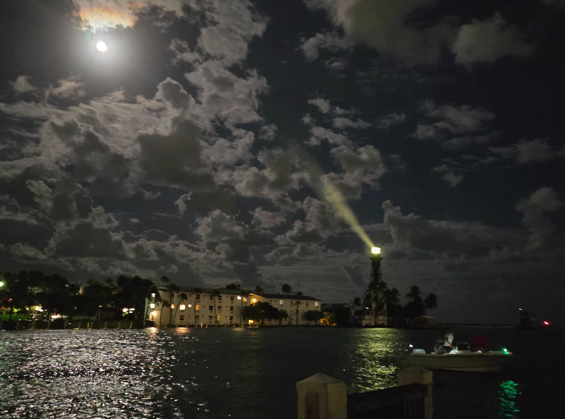 A full moon is photographed shining in a cloudy night sky, while a light house directs its beam skyward on the water's edge.
