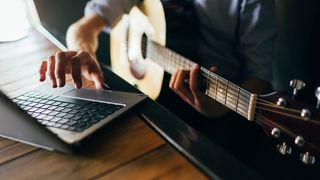 Person plays an acoustic guitar with a laptop computer in front of them on a desk