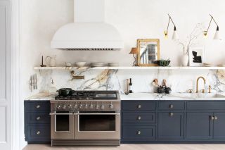 Image of a white kitchen with navy cabinetry and white marble countertops and backsplash. There is a wall-to-wall floating shelf and a large white cooker hood.