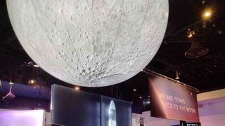huge moon is attached to the ceiling of a museum. to the right is a big display saying "We are going back to the moon"