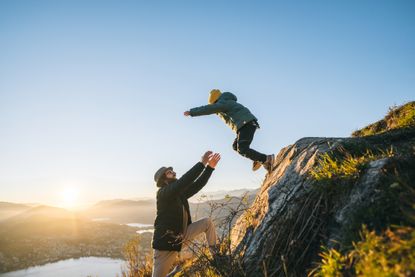 A son jumps into fathers arms on mountain top above lake at sunset. The jump exemplifies trust.