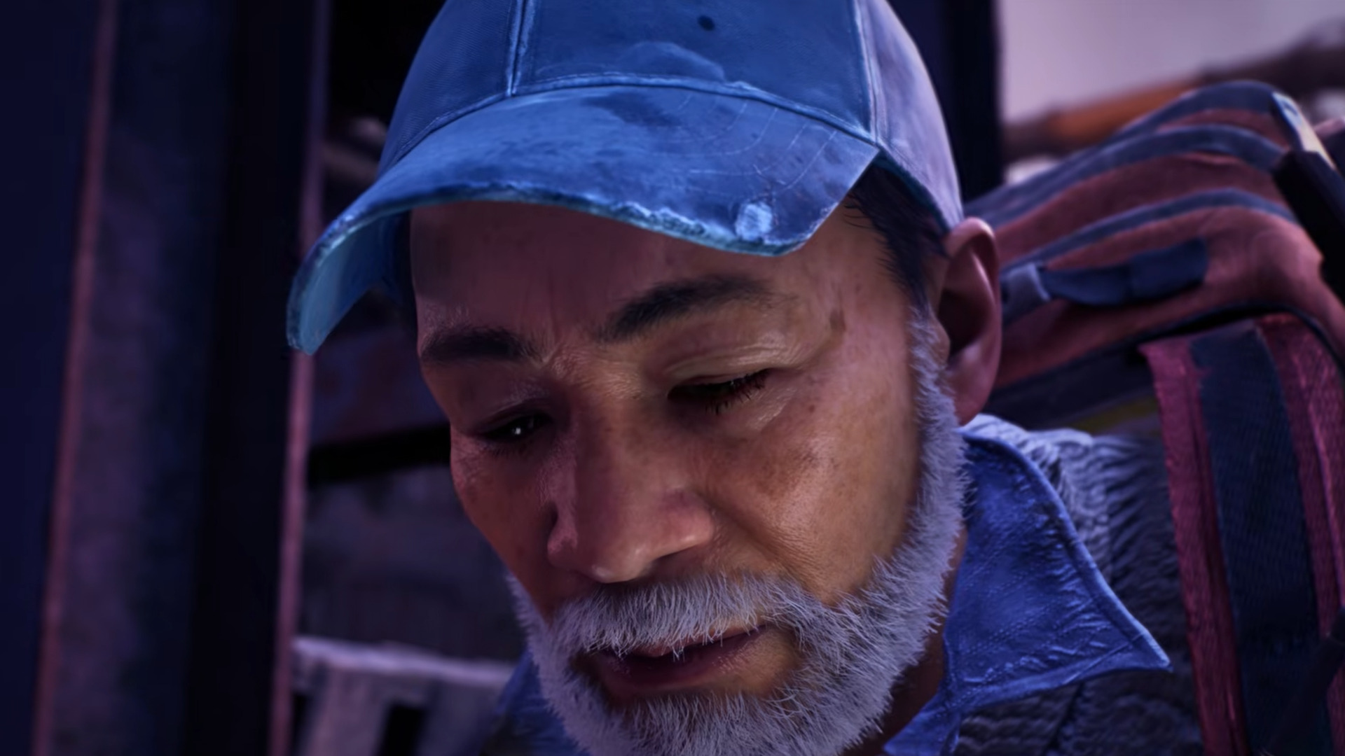 Older man with gray bears and jean hat looks down at ground with a grave expression